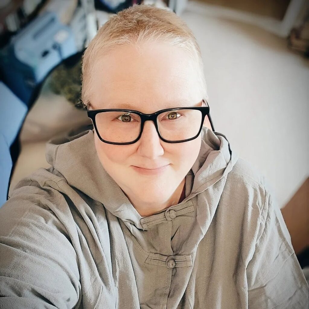 Photo shows a woman with very short blonde-looking hair wearing black-framed glasses and a grey, hooded tunic smiling at the camera.